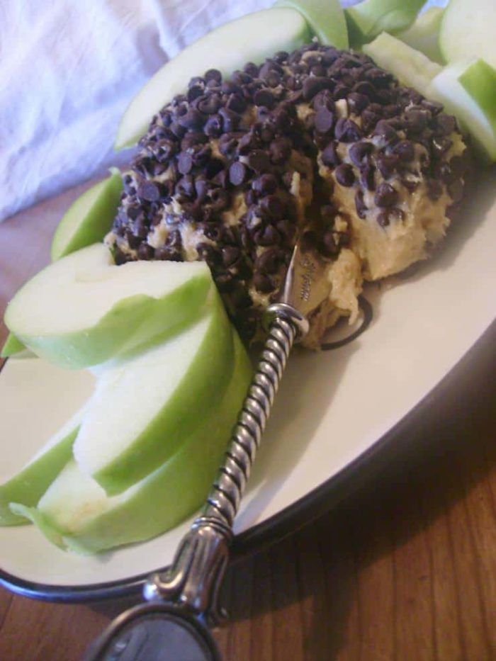 Image shows a peanut butter cheeseball covered in chocolate chips on a plate with apples