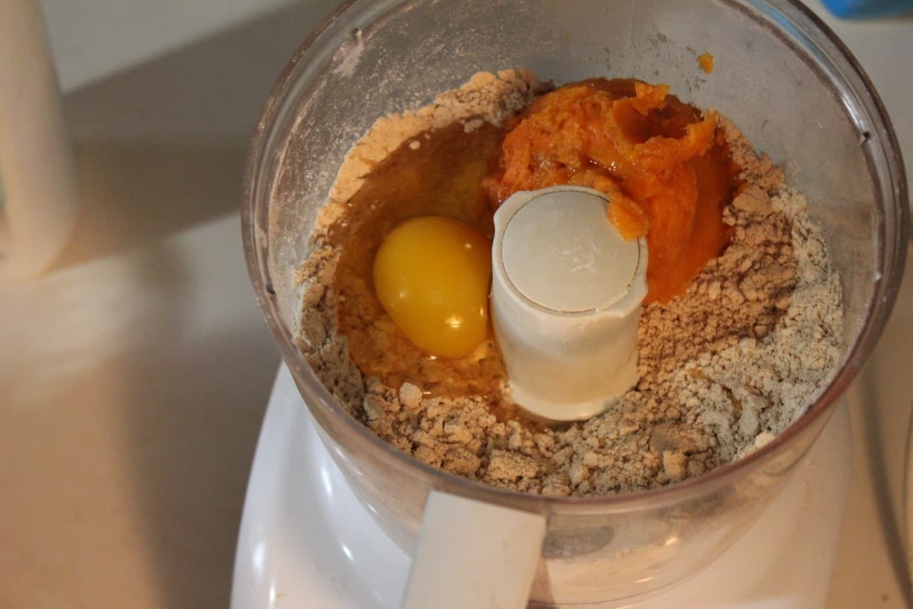 Image shows a food processer, photographer from above, with gluten free flour, egg and sweet potato ready to be blended.
