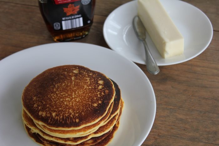 Photo shows a stack of pancakes on a white plate next to some butter