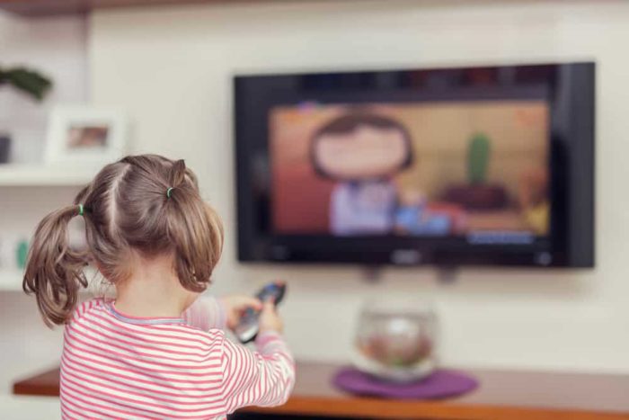 Photo shows a young girl with pigtails holding a remote and watching TV