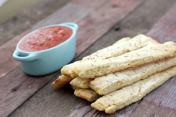 Photo shows a stack of breadsticks on a table with a dish of marinara sauce