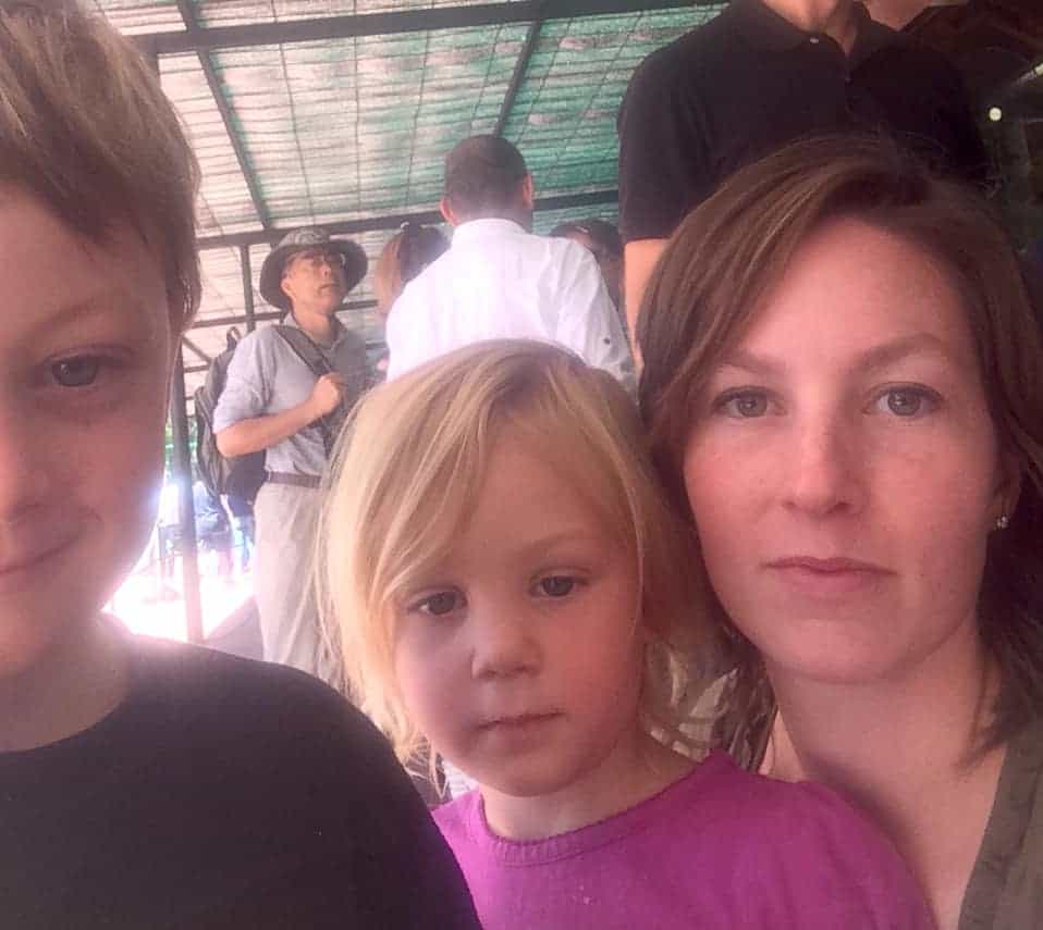 Image shows a woman with brown hair, holding a young blonde girl in her lap, with a young boy in the forefront of the photo.