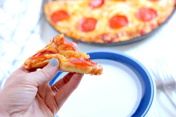 Image shows a hand holding a slice of pepperoni pizza over a white and blue plate. In the background is the rest of the cauliflower crust pizza