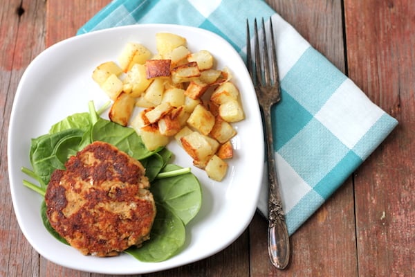 Image shows a salmon patty on a bed of lettuce with potatoes, next to a napkin with a fork