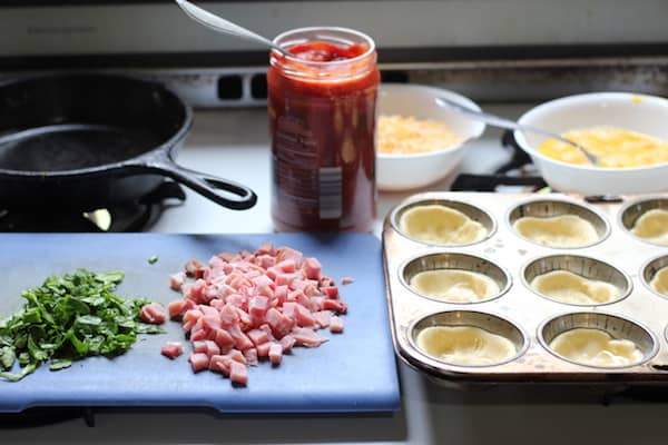 Image shows a cutting board of ingredients next to an iron skillet, and muffin tin of pizza crusts