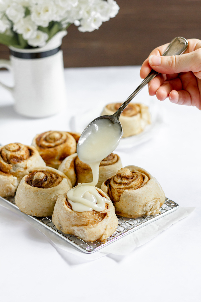 Image shows a hand drizzling icing over cinnamon rolls