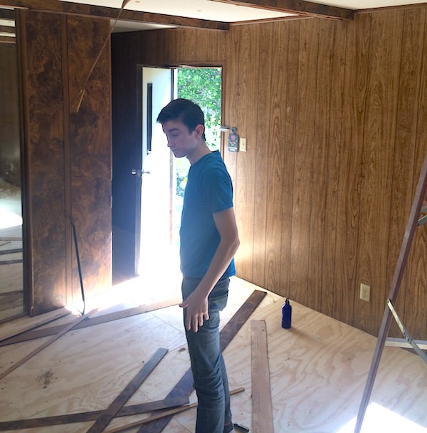 Image depicts a young man in a wood paneled room near a door