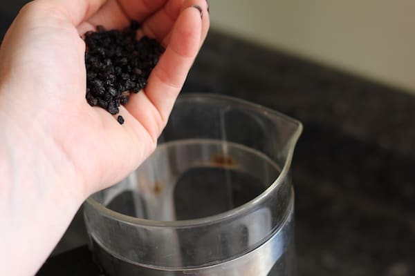 Photo shows a hand holding elderberries over a tea carafe