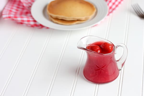 Image shows a small glass vessel with strawberry sauce next to a plate of pancakes