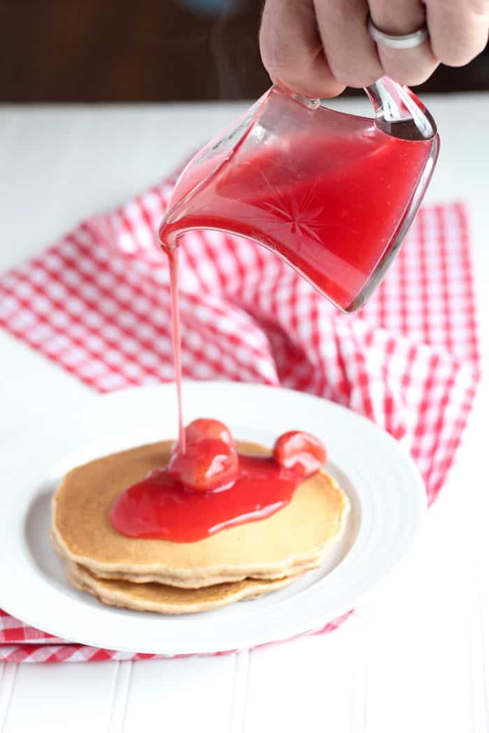 Image shows a hand pouring strawberry syrup over a plate of pancakes