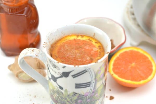 Photo shows a white mug with a clock on it filled with orange detox tea in the foreground, with a slice of orange floating in it.Next to the mug is a half an orange sliced in half, and a container of honey.
