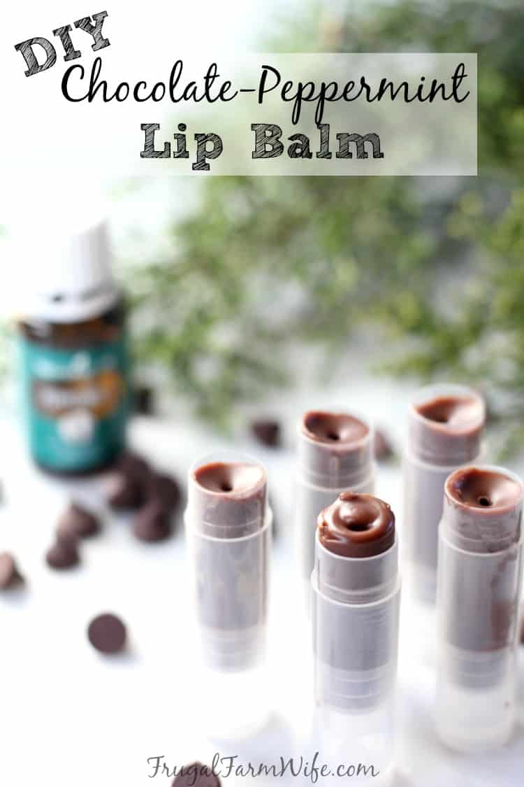 Image shows a close up of several tubes of chocolate mint lip balm, standing on their ends. In the background are scattered chocolate chips, and some mint leaves. Text overlay reads "DIY Chocolate-Peppermint Lip Balm"