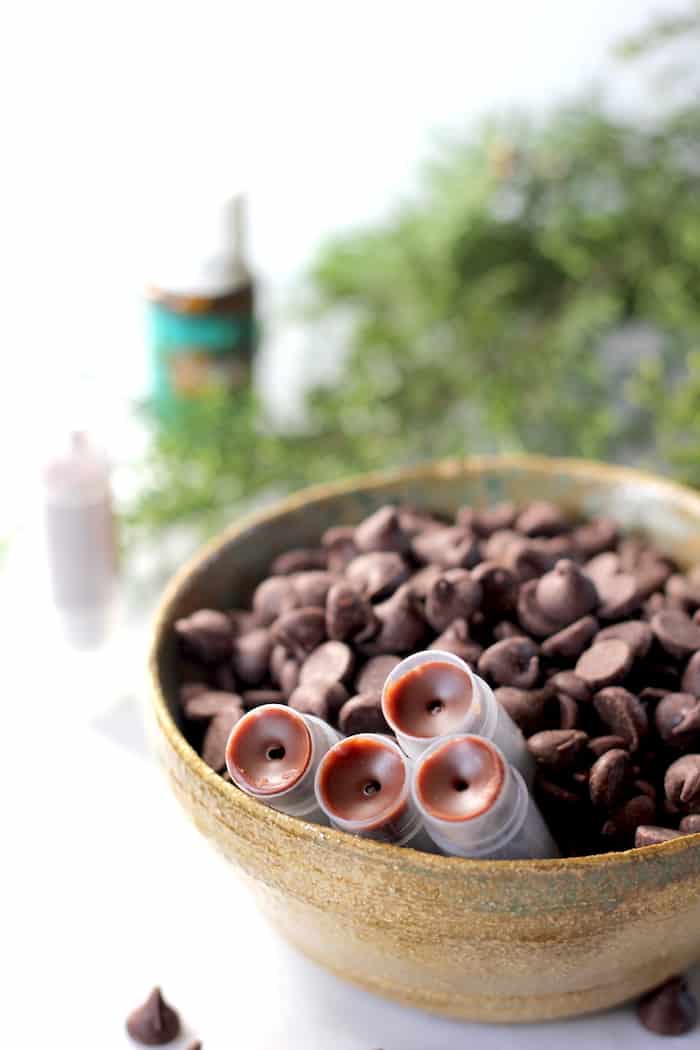 Image shows a ceramic bowl full of chocolate chips, with 4 tubes of chocolate peppermint lip blam in it. 