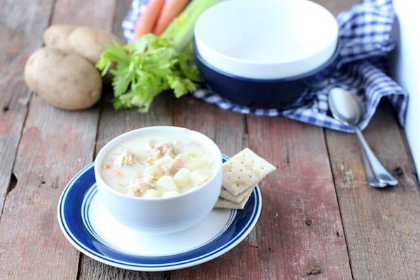 Photo shows a bowl of clam chowder soup on a table with empty bowls, some potatoes, carrots and celery nearby.