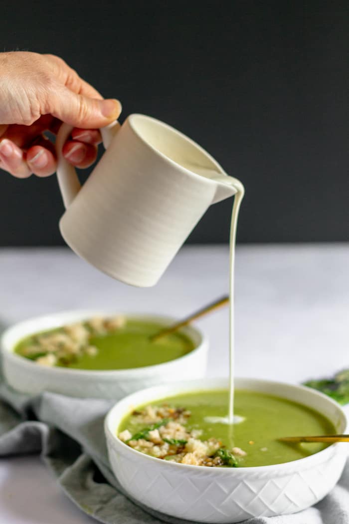 Image shows a hand pouring cream into a bowl of cream of asparagus soup