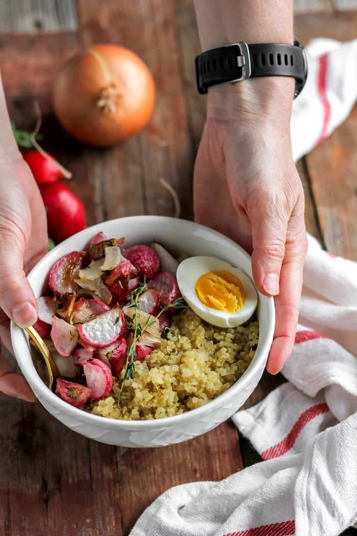 Photo shows two hands holding a bowl of roasted radishes with quinoa and a hard boiled egg