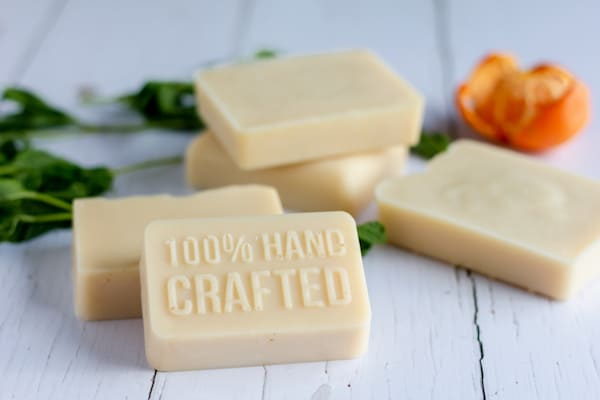 Photo shows five bars of cream colored goat milk on a white table. The front bar reads "100% Hand Crafted." Spearmint leaves and an orange sit in the background.