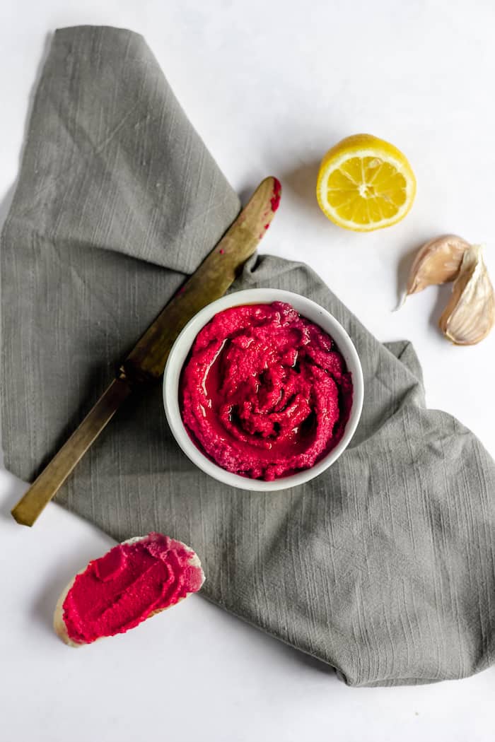 Image shows a bowl of red beet hummus next to a knife, on a grey napkin