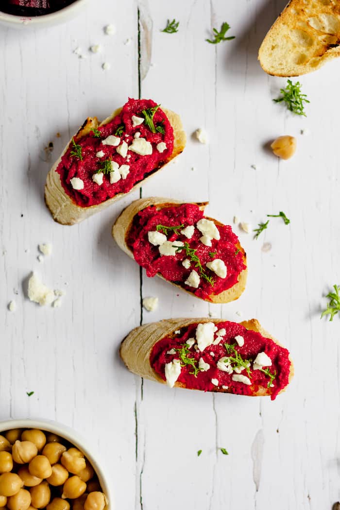 Image shows several slices of toasted baguette, with roasted red beet hummus and pieces of feta on top