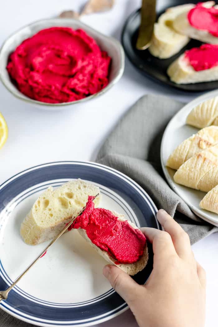 Image shows a hand spreading hummus with beets on bread
