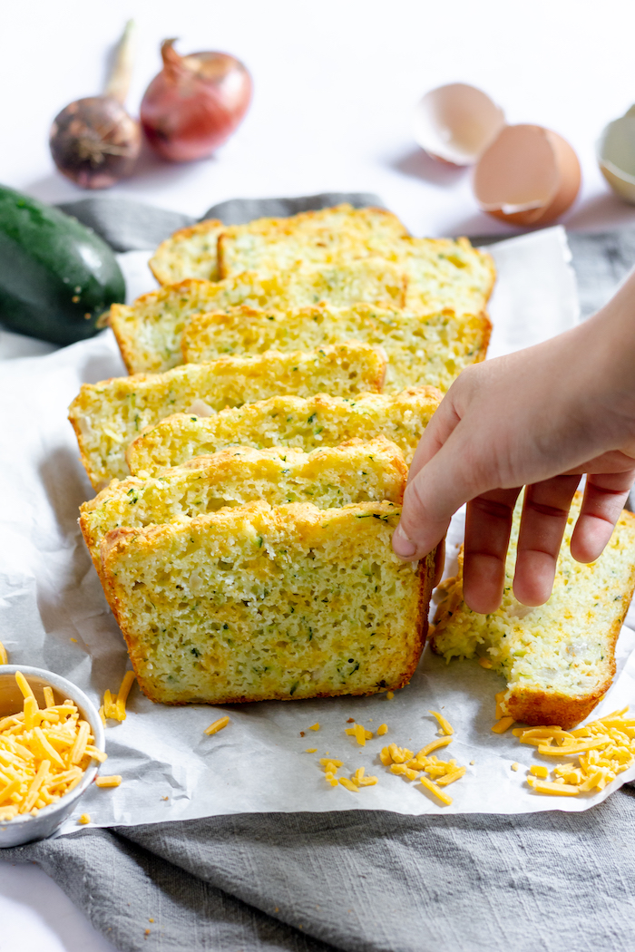 Image shows a hand taking a slice of cheesy zucchini bread