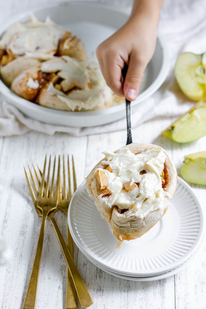 Image shows a hand holding an apple cinnamon roll over a white plate