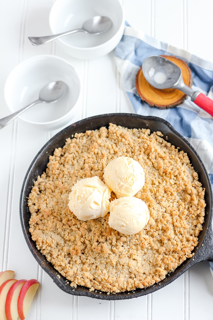 Image shows an apple crisp in a skillet next to two empty bowls and an ice cream scoop