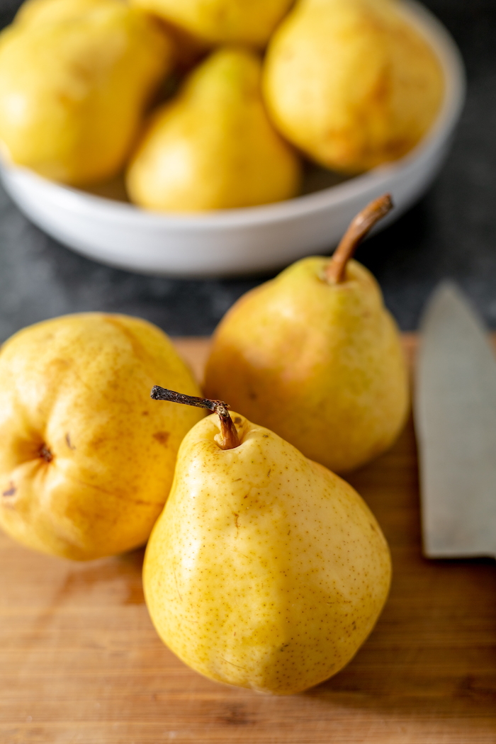 Image shows a close up of three yellow Bartlett pear on a wood cutting board with a knife sitting next to them. Behind the pears is a white bowl of more pears.