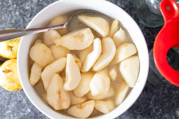 Photo, taken from above, shows a white bowl of sliced pears in lemon water, with a spoon. Next to the bowl sits two uncut pears.