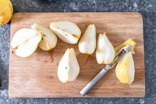 Photo, taken from above, shows several pears sliced into quarters, on a cutting board. With the pears sits a peeler.
