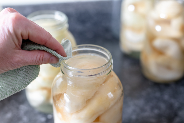 Image shows a hand holding a washcloth drying the edge of the mason jar full of pears and syrup. Several more jars sit in the background.