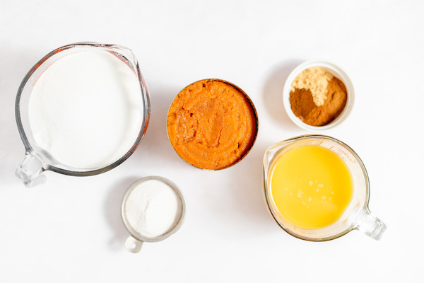 Photo, taken from above, shows the ingredients needed to make pumpkin jam- a glass measuring cup of sugar, a jar of pumpkin puree, a measuring cup of orange juice, and two small dishes of spices