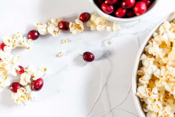 Image shows a string of popcorn garland on a table, with a bowl of popcorn and cranberries to make the garland. 